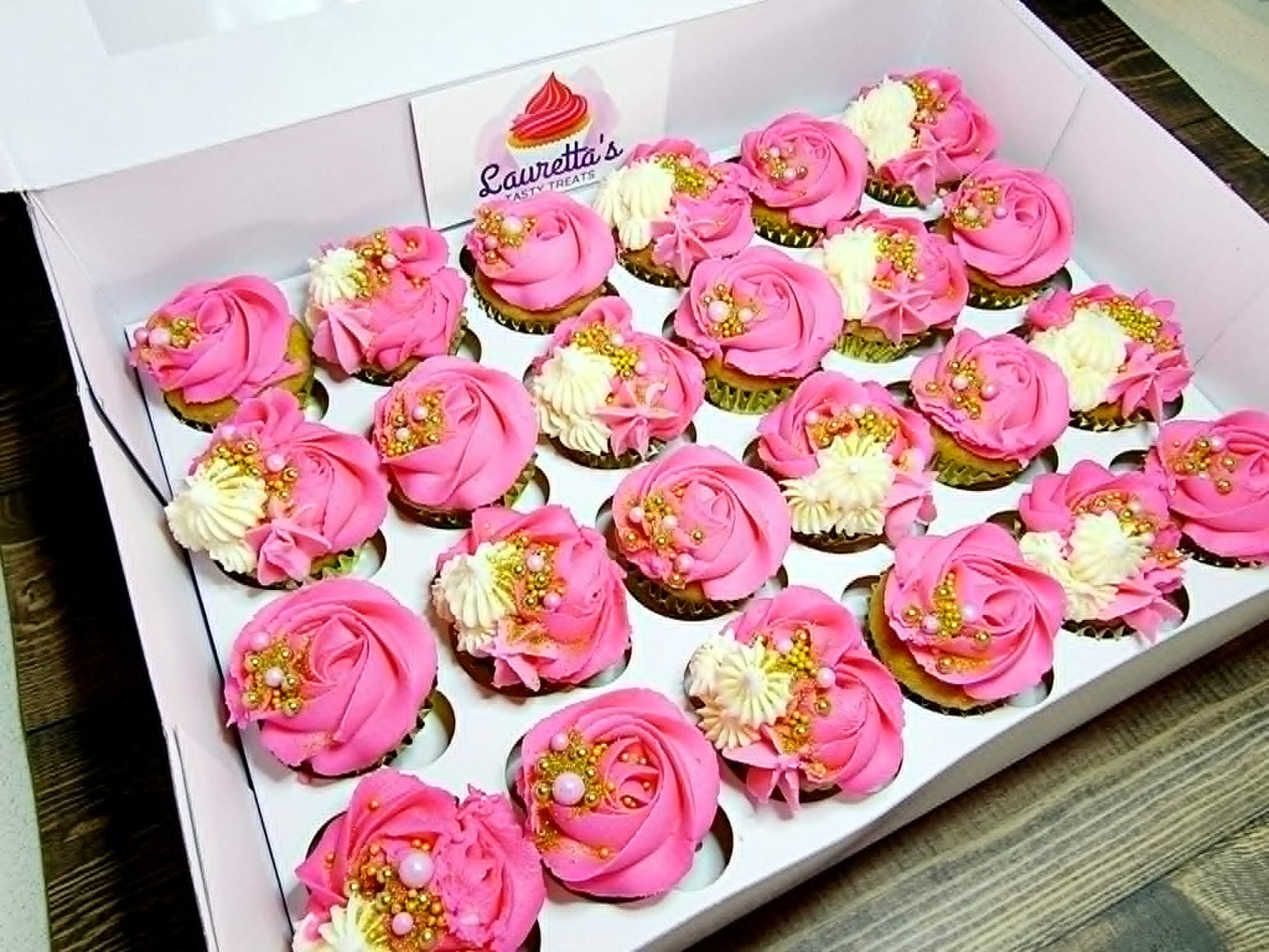 Box of cupcakes with pink and white frosting, decorated like flowers, with edible gold decorations.